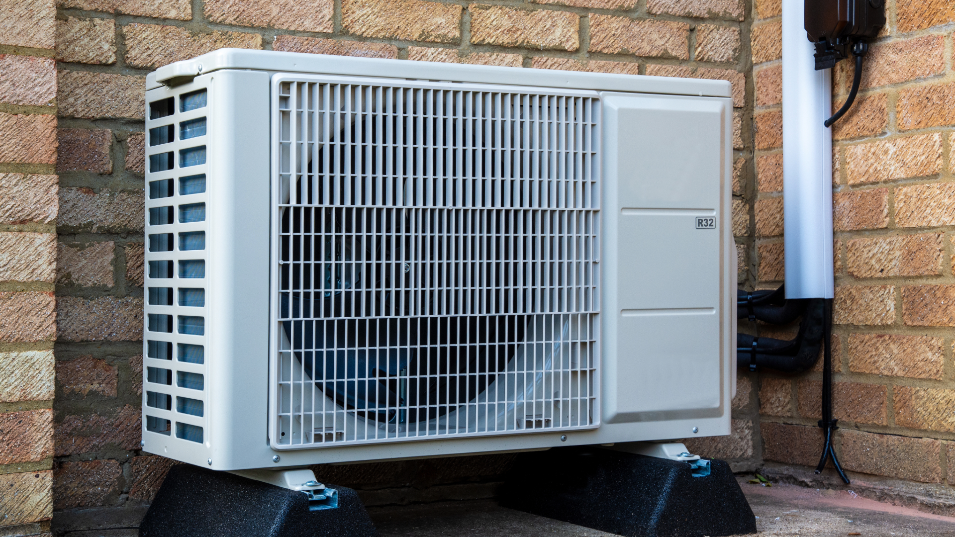 Air source heat pump unit mounted on rubber feet against a brick house wall.