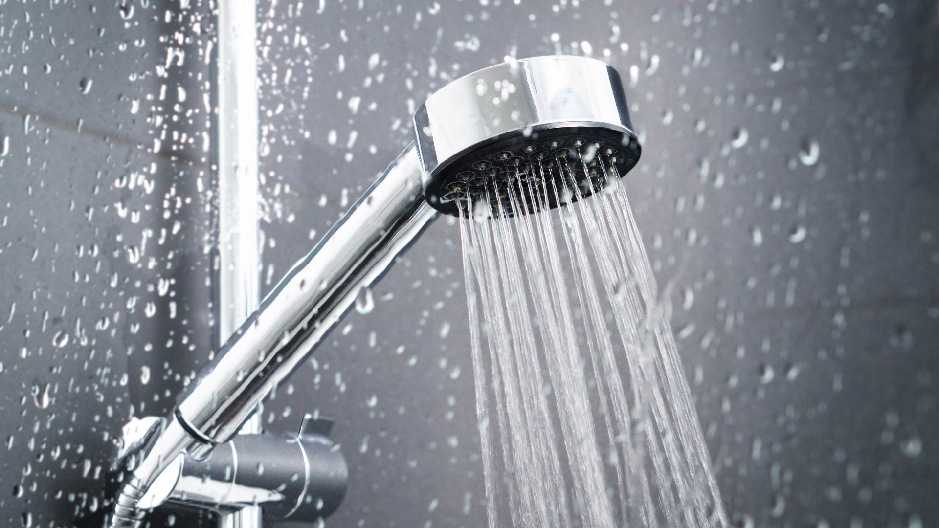 Chrome handheld showerhead spraying water against a dark, water-splashed tiled wall.