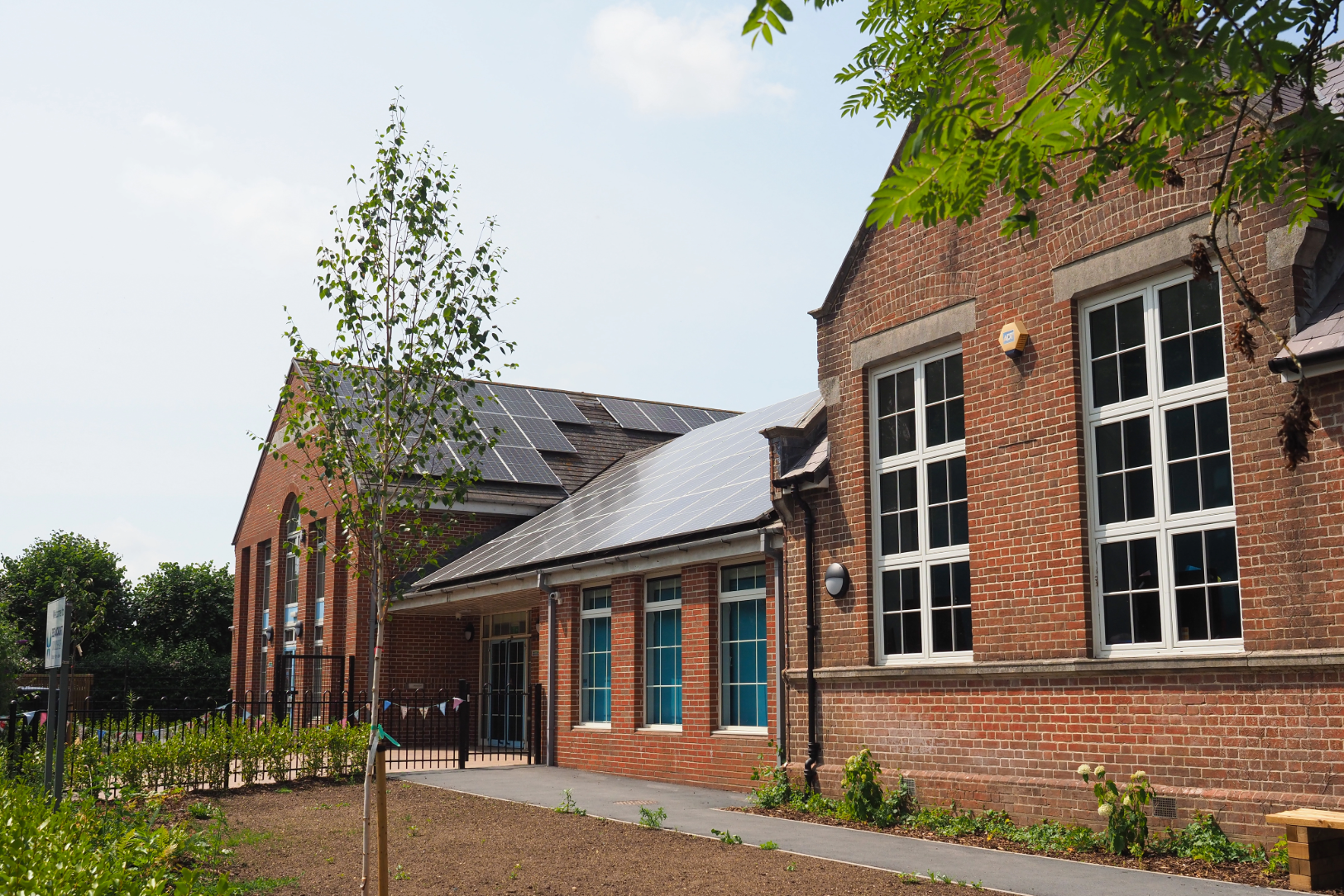 Traditional red-brick school building with solar panels installed across its pitched roofs, with a young tree and iron railings in the foreground.