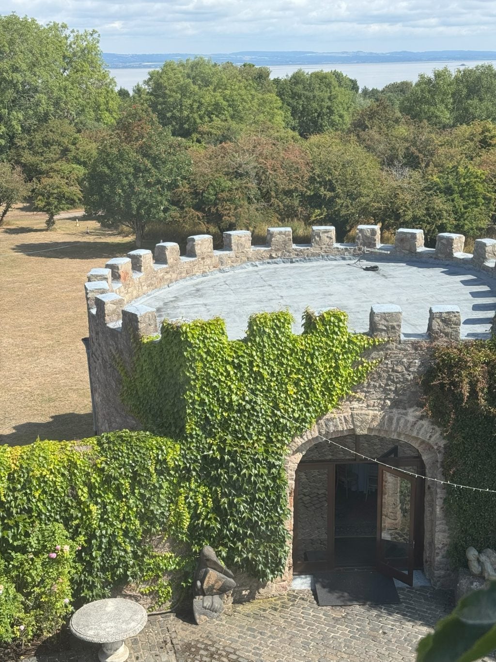 A scenic view from a high vantage point at Walton Castle, looking down onto a stone turret and ivy-covered archway, with the Bristol Channel visible in the distance.