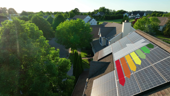An aerial view of a residential neighbourhood with houses and green trees. In the foreground, a house roof covered in solar panels is prominently featured. Overlaid on the solar panels is a large graphical representation of the standard energy performance rating chart, with colourful arrows ranging from red (G) to green (A), symbolising EPC reform.