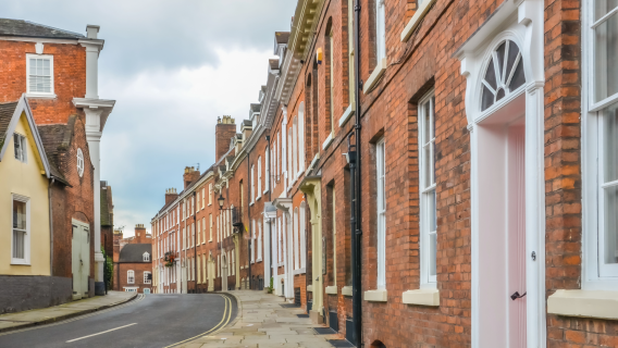 A winding street in Shrewsbury, Shropshire, lined with historic red-brick terraced houses typical of the hard-to-treat housing stock targeted by the Warm Homes: Local Grant. The three-storey properties feature sash windows and solid walls, illustrating the technical challenges of delivering PAS 2035 compliant retrofit and energy efficiency upgrades in heritage areas.