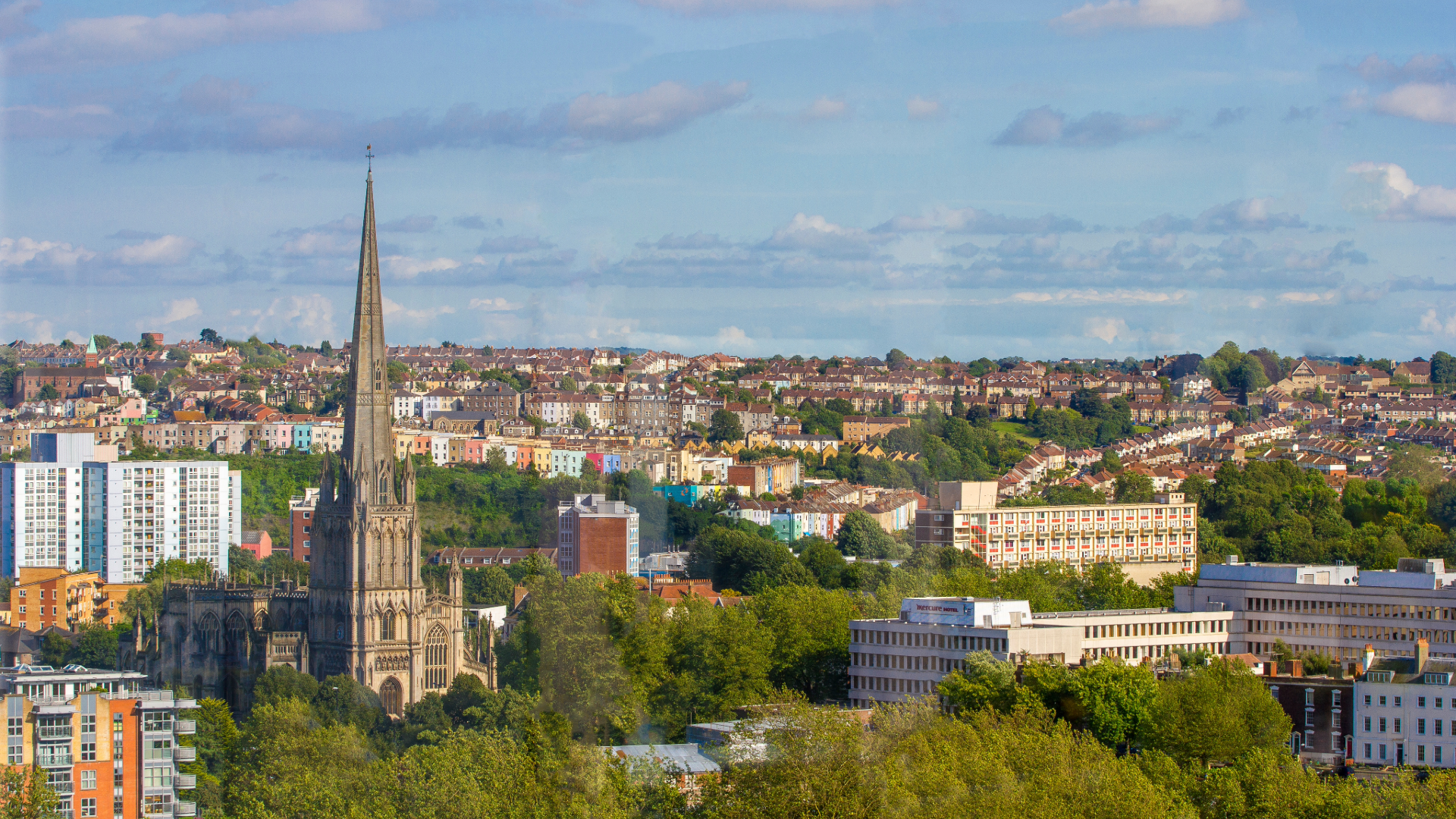 Aerial view of Bristol cityscape with a Gothic church spire in the foreground and colourful terraced housing across the hillside
