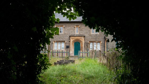 Exterior of the Old Frenchay Primary School, a stone former school building with a Gothic-arched teal door, seen through overgrown grass and a low picket fence.