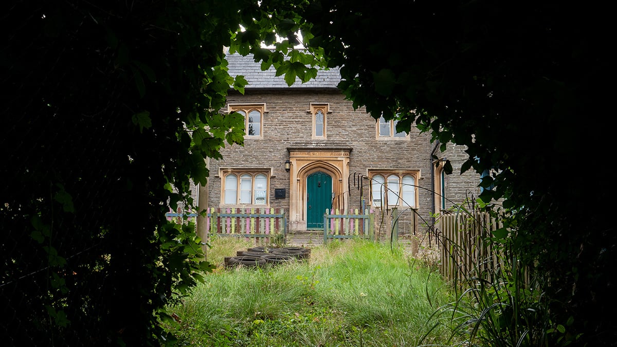 Exterior of the Old Frenchay Primary School, a stone former school building with a Gothic-arched teal door, seen through overgrown grass and a low picket fence.