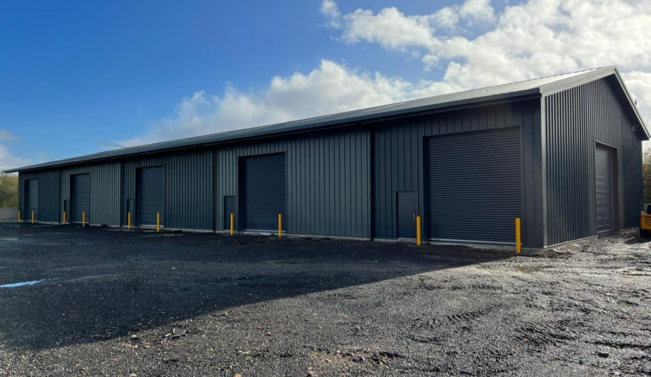 The newly completed industrial unit at Winkleigh Airfield, Devon, with grey metal cladding and five roller shutter doors on a gravel yard.