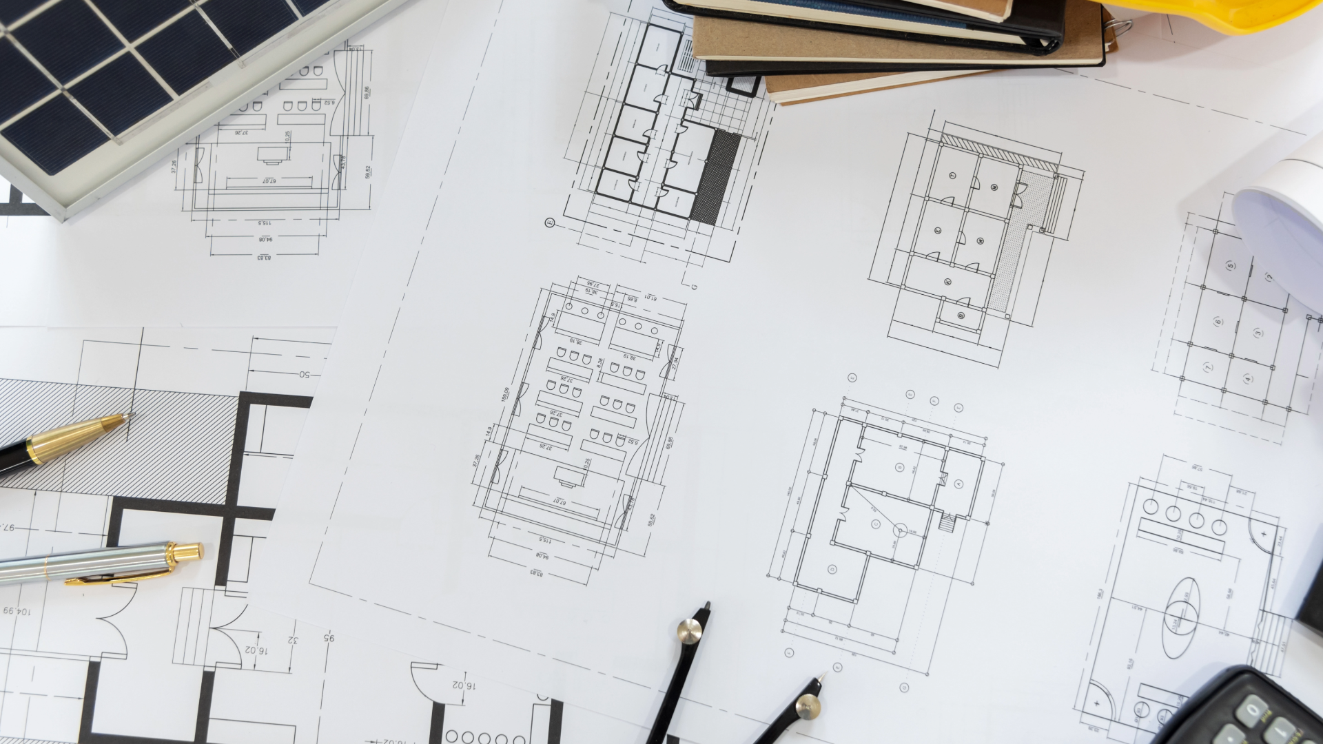 Overhead view of architectural floor plans spread across a desk, with two fountain pens, a compass, a solar panel, a stack of notebooks, and a calculator.